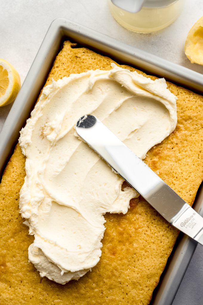 Someone is using an offset spatula to spread lemon buttercream frosting onto a lemon sheet cake.