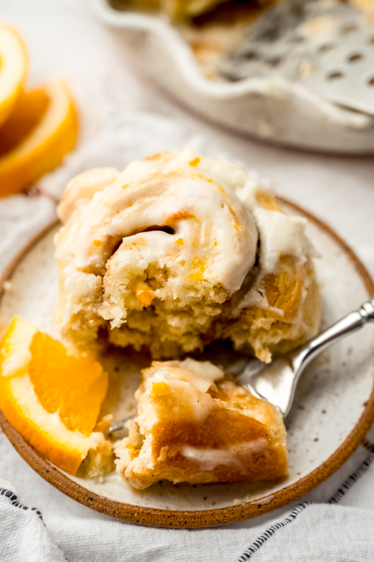 An orange cinnamon roll sitting on a plate with a fork that has a bite on it and a small wedge of an orange off to the side.