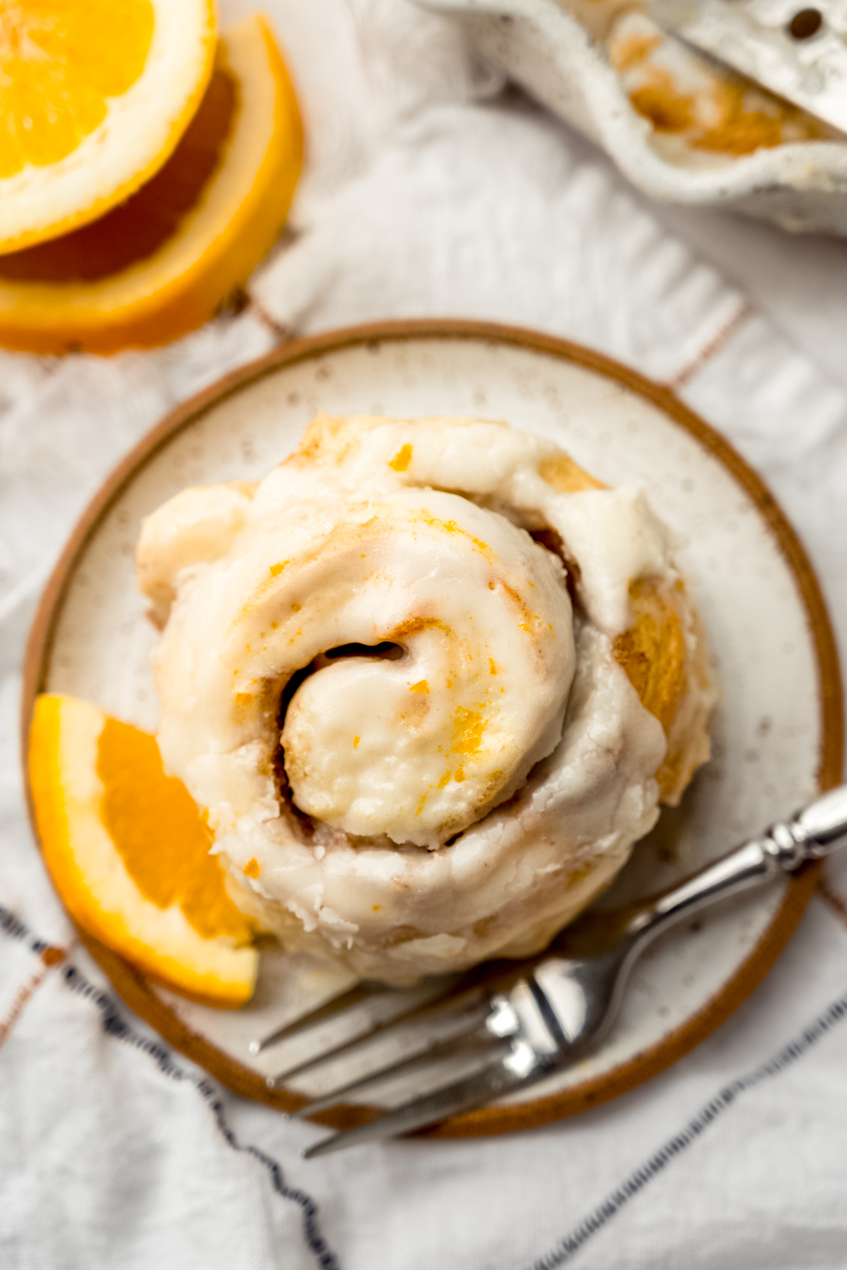 Aerial photo of an orange cinnamon roll sitting on a plate with a fork and a small wedge of an orange.