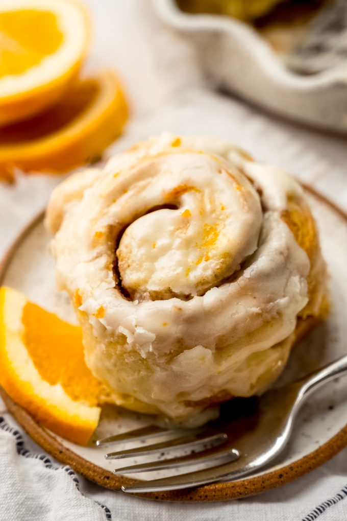 An orange cinnamon roll sitting on a plate with a fork and a small wedge of an orange.