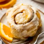 An orange cinnamon roll sitting on a plate with a fork and a small wedge of an orange.