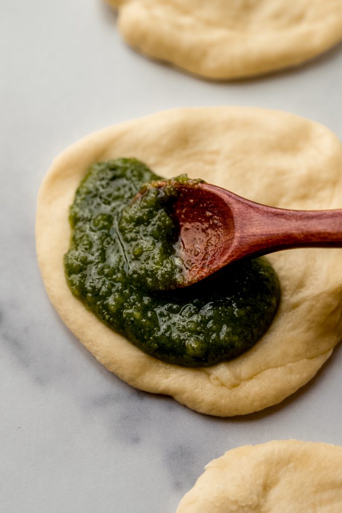 Someone is using a small spoon to put pesto onto a circle of yeasted dough to make pull-apart bread.