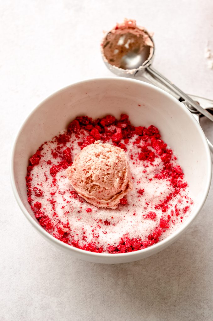 A ball of strawberry sugar cookie dough in a bowl of sugar and freeze-dried strawberry coating.