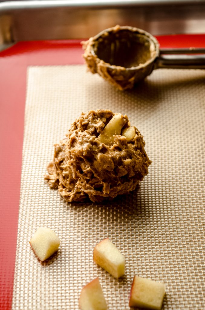 An apple oatmeal cookie dough ball on a baking sheet with a cookie scoop and apple slices around it.