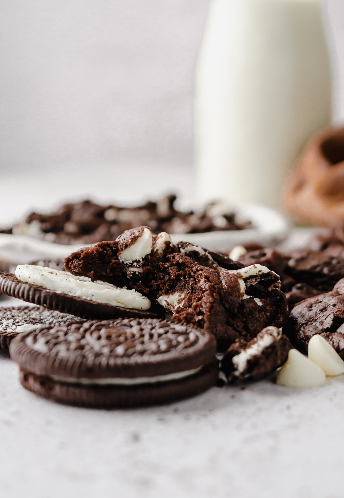 A chocolate cookies and cream cookie with a bite taken out of it sitting on a surface with a bottle of milk in the background.