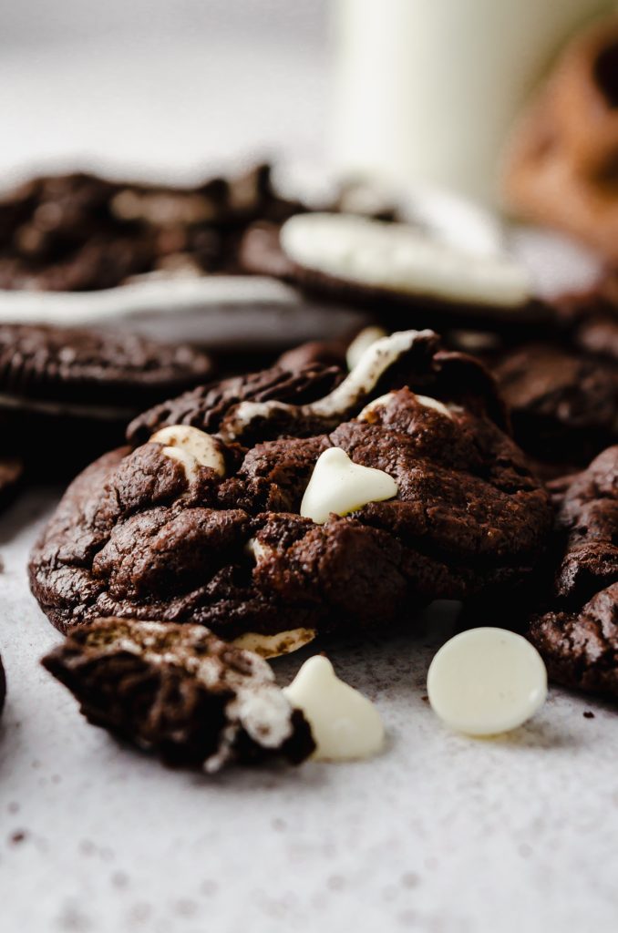 A chocolate cookies and cream cookie on a surface with white chocolate chips and Oreo chunks around it.