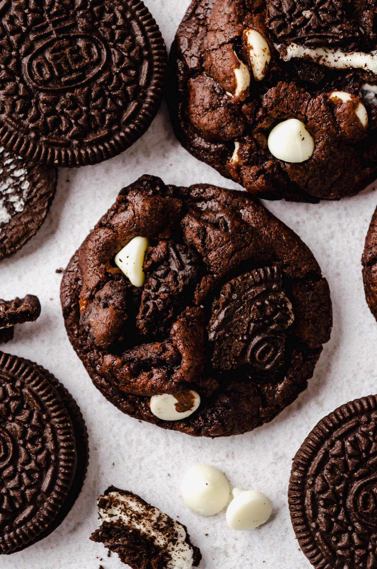 A chocolate cookies and cream cookie on a surface with white chocolate chips and Oreo chunks around it.