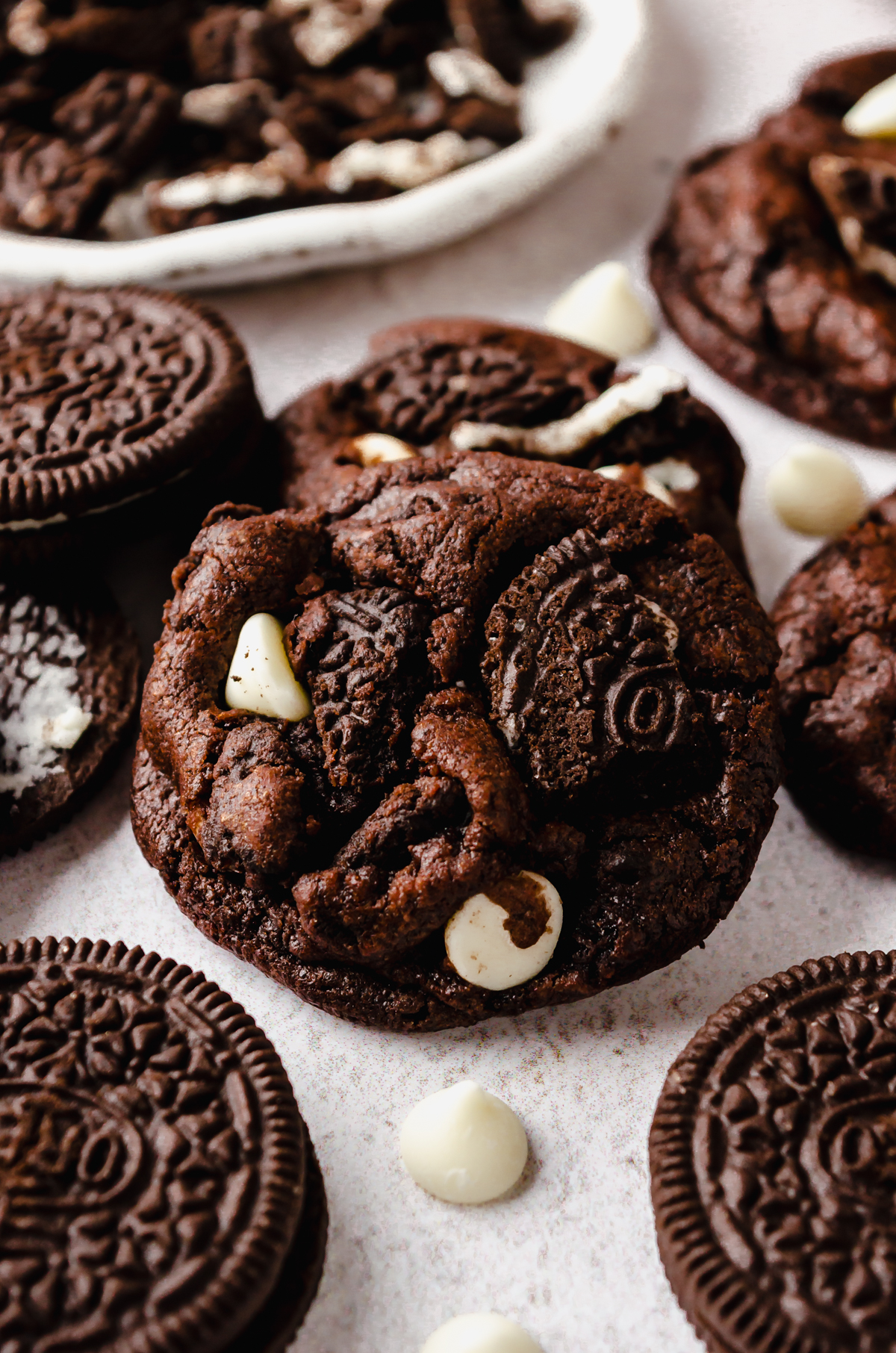 A chocolate cookies and cream cookie on a surface with white chocolate chips and Oreo chunks around it.