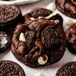 A chocolate cookies and cream cookie on a surface with white chocolate chips and Oreo chunks around it.