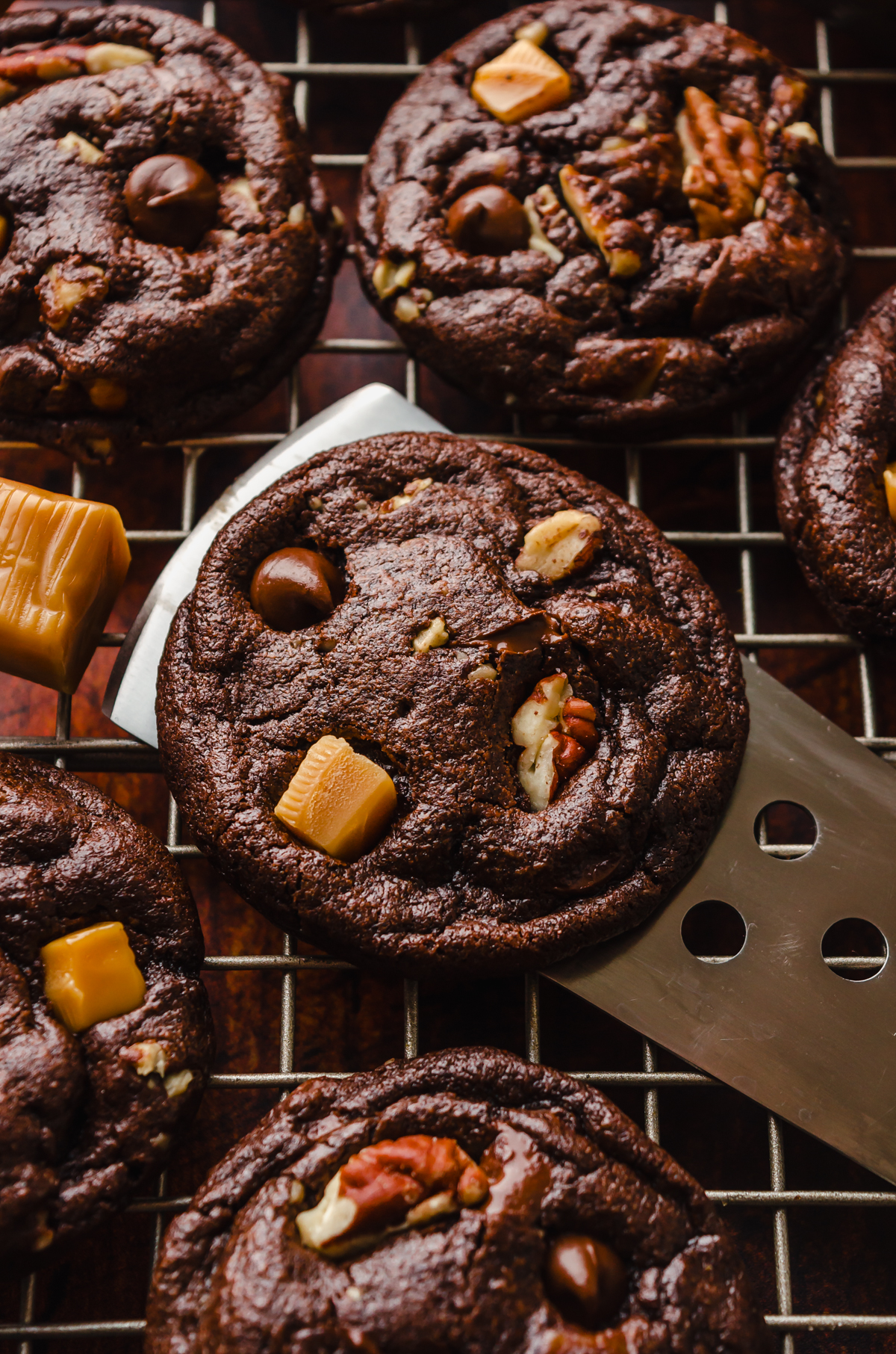 Someone is lifting a chocolate turtle cookie off of a cooling rack with a spatula.