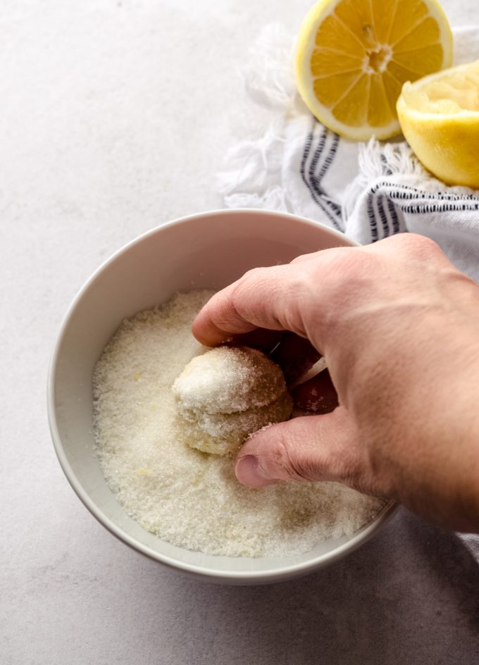 Someone is using their hands to roll a ball of lemon sugar cookie dough in a bowl of lemon sugar.