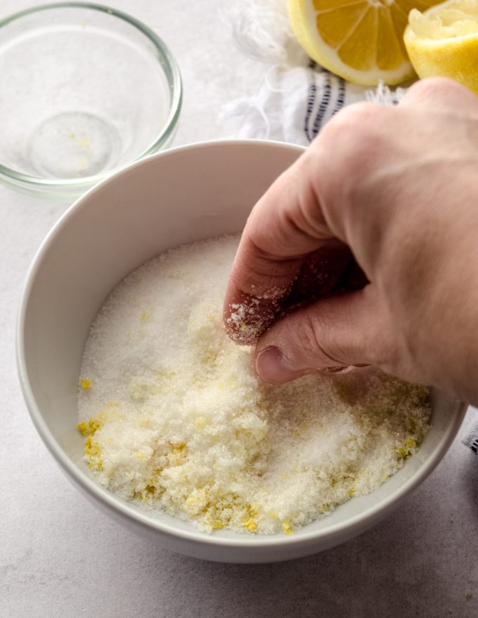 Someone is using their fingers to rub lemon zest and sugar together to make a lemon sugar coating for lemon sugar cookies.