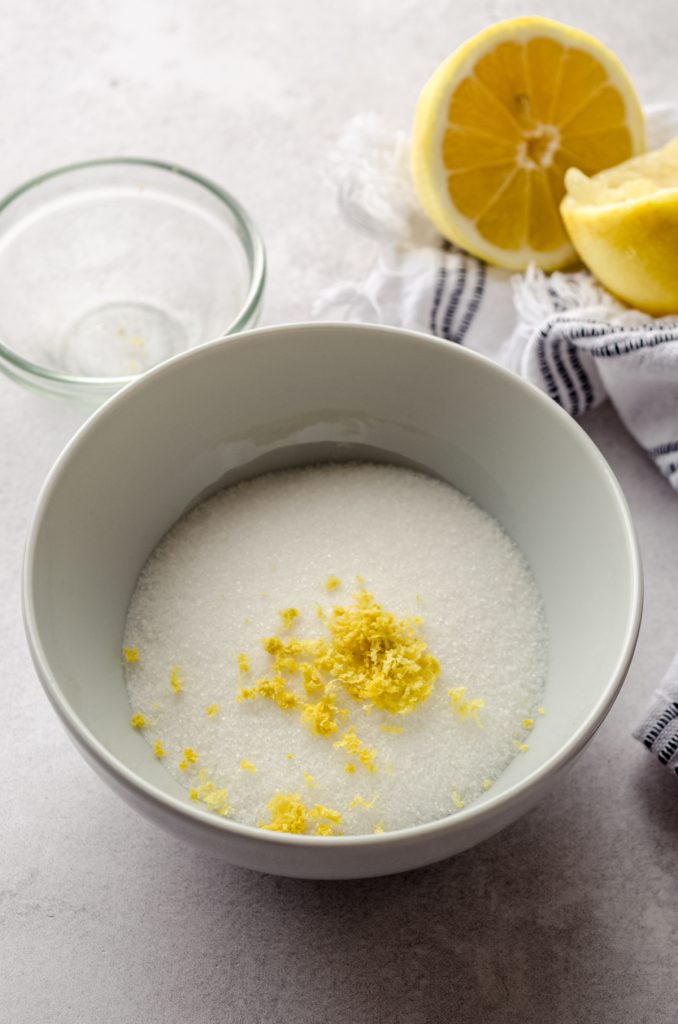 A bowl of sugar and lemon zest with lemons in the background.