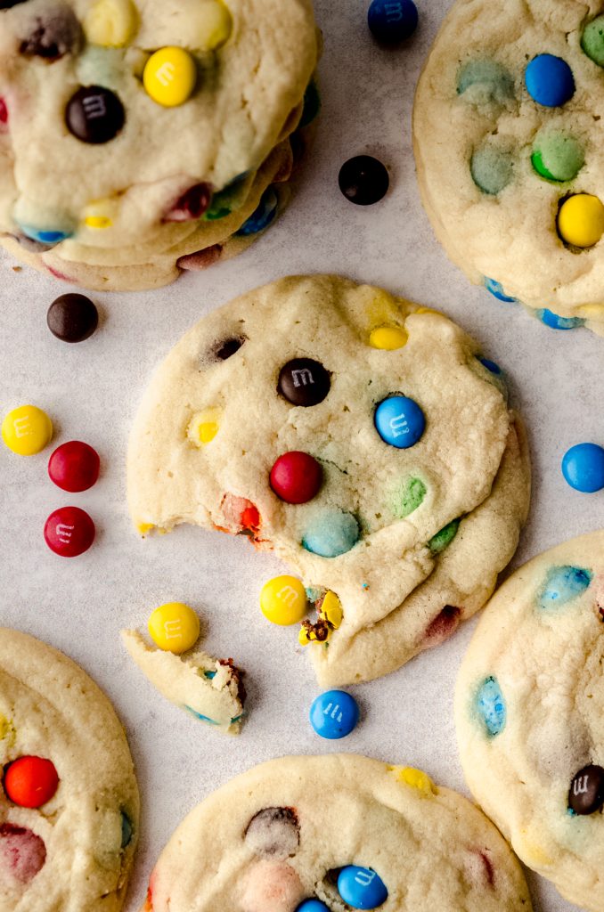 Aerial photo of a m&m sugar cookie on a surface with a bite taken out of it.