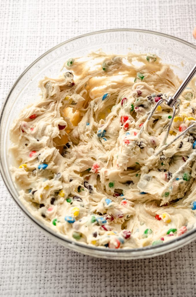 A bowl of m&m sugar cookie dough with electric beaters in it.