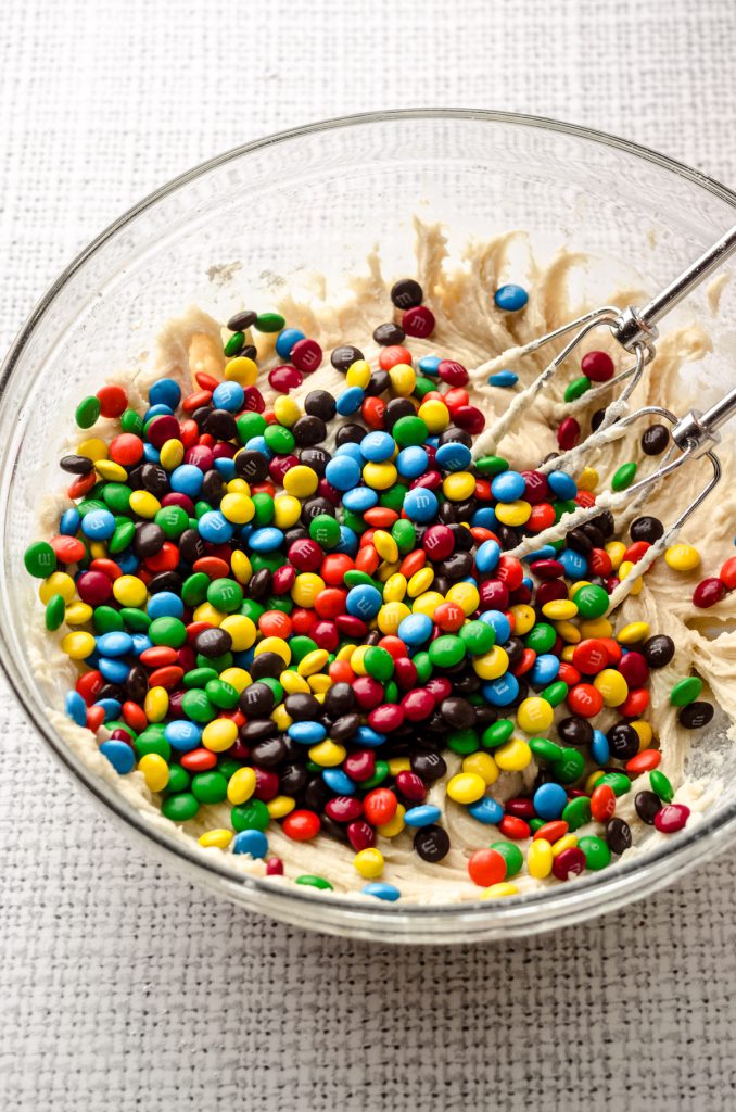 A bowl of m&m sugar cookie dough with electric beaters in it.