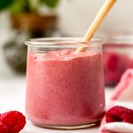 A jar of raspberry curd with a spoon in it and plants in the background and raspberries in the foreground.