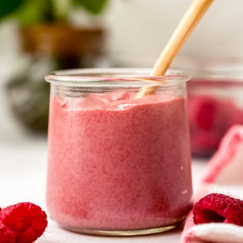A jar of raspberry curd with a spoon in it and plants in the background and raspberries in the foreground.