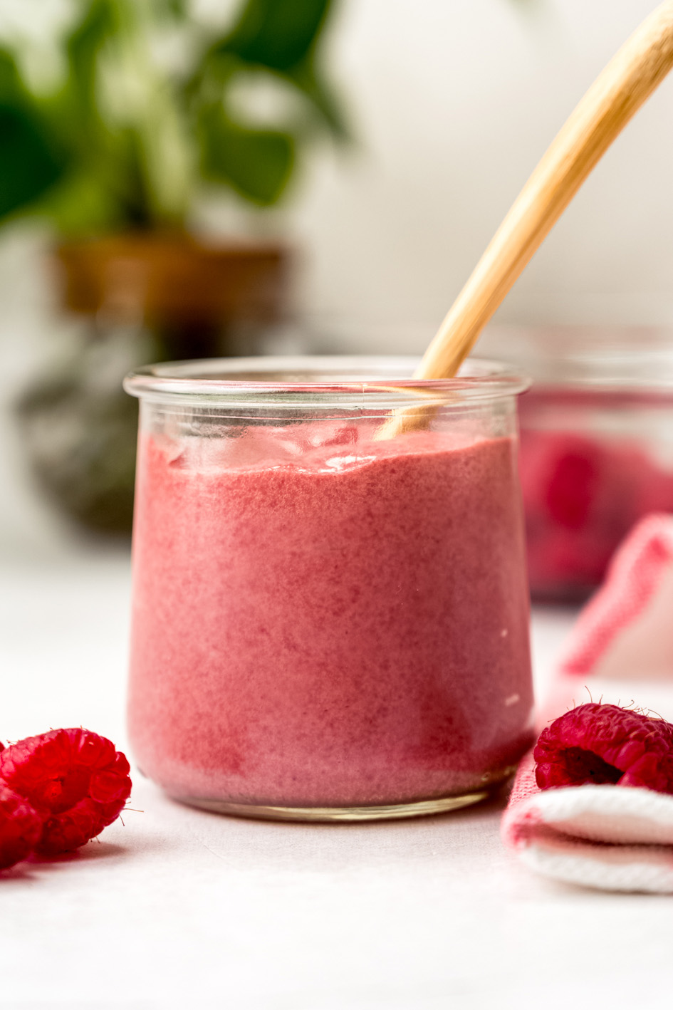 A jar of raspberry curd with a spoon in it and plants in the background and raspberries in the foreground.