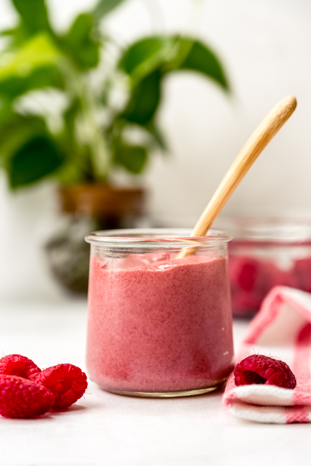 A jar of raspberry curd with a spoon in it and plants in the background and raspberries in the foreground.