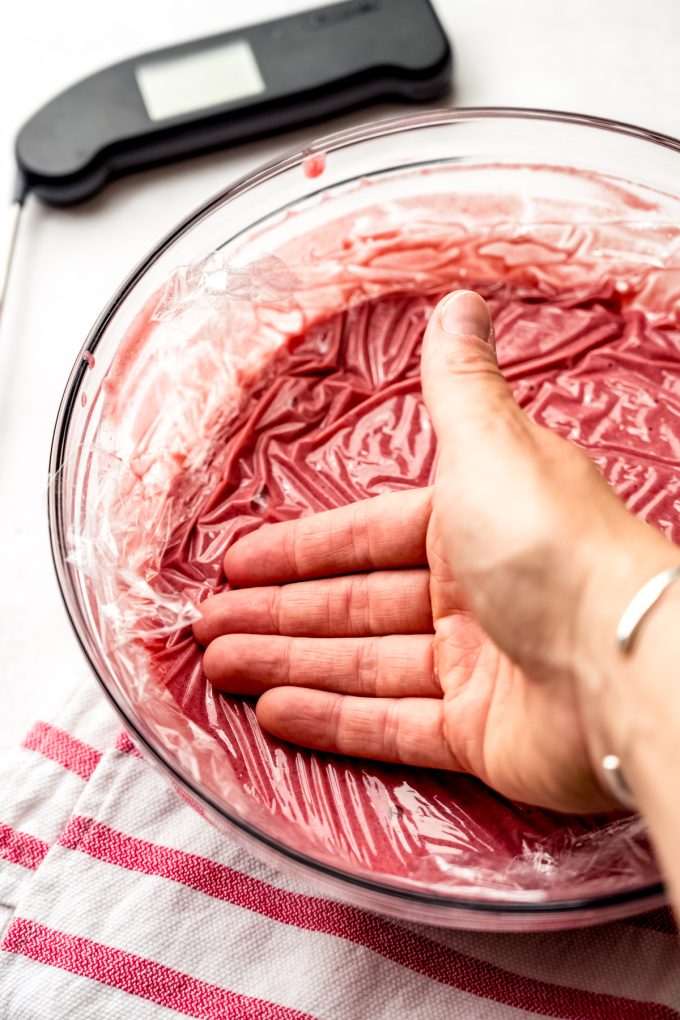 Someone is pressing plastic wrap onto a bowl of warm raspberry curd.