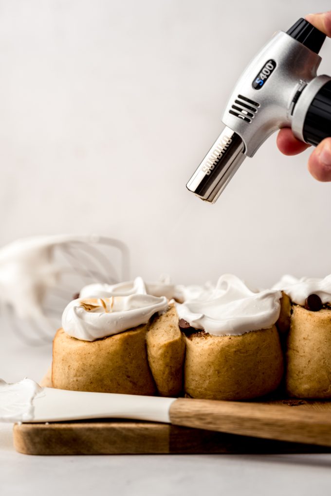Someone is using a kitchen torch to brown the marshmallow meringue on top of s'mores cinnamon rolls.