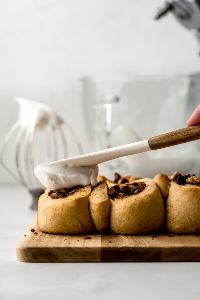 Someone is using a spatula to spread marshmallow meringue onto s'mores cinnamon rolls.