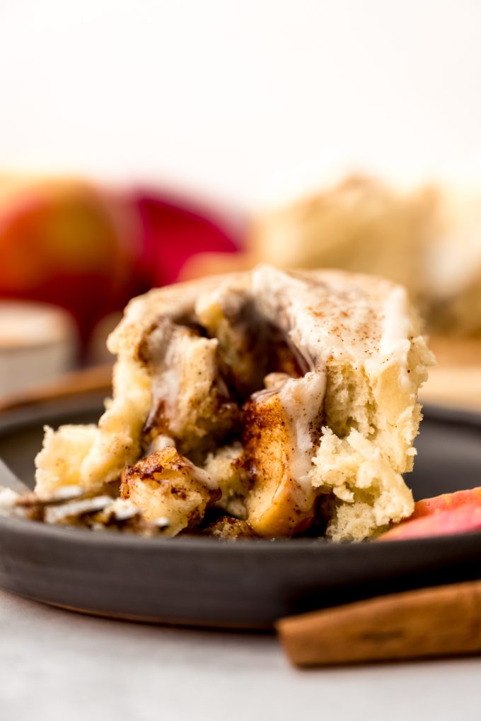 An apple cinnamon roll on a plate with a fork and apple slices and cinnamon sticks in the foreground.