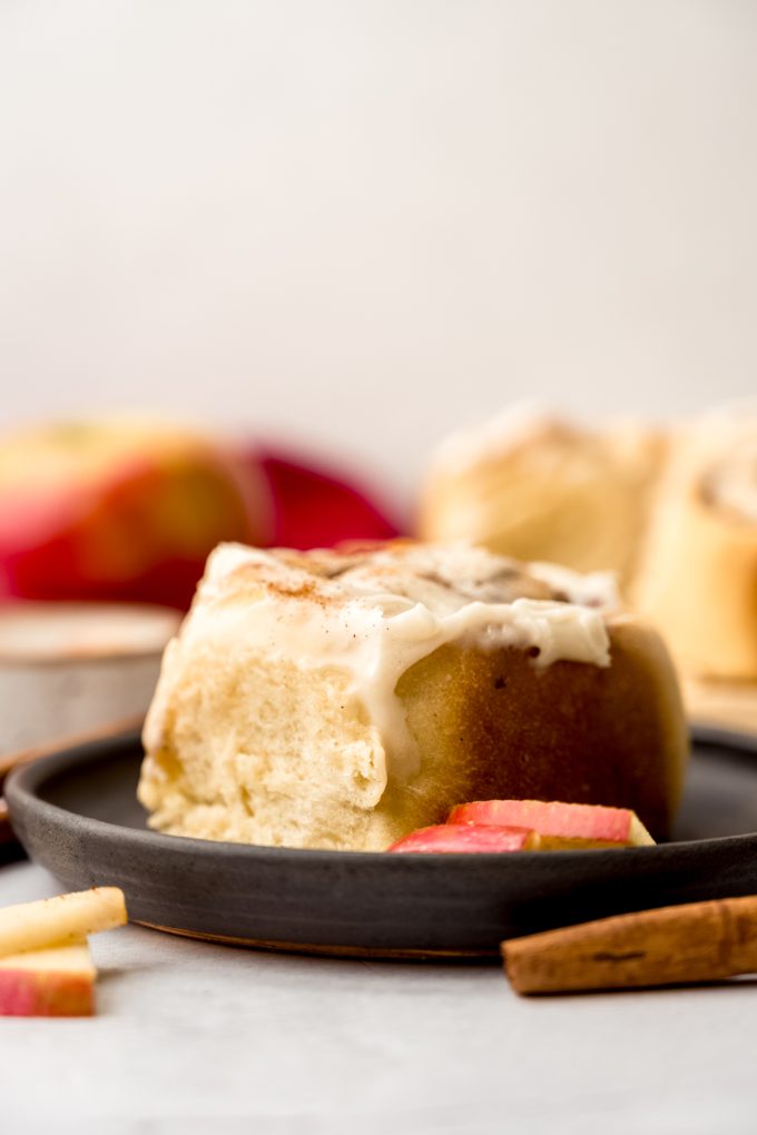 An apple cinnamon roll on a plate with apple slices and cinnamon sticks in the foreground.