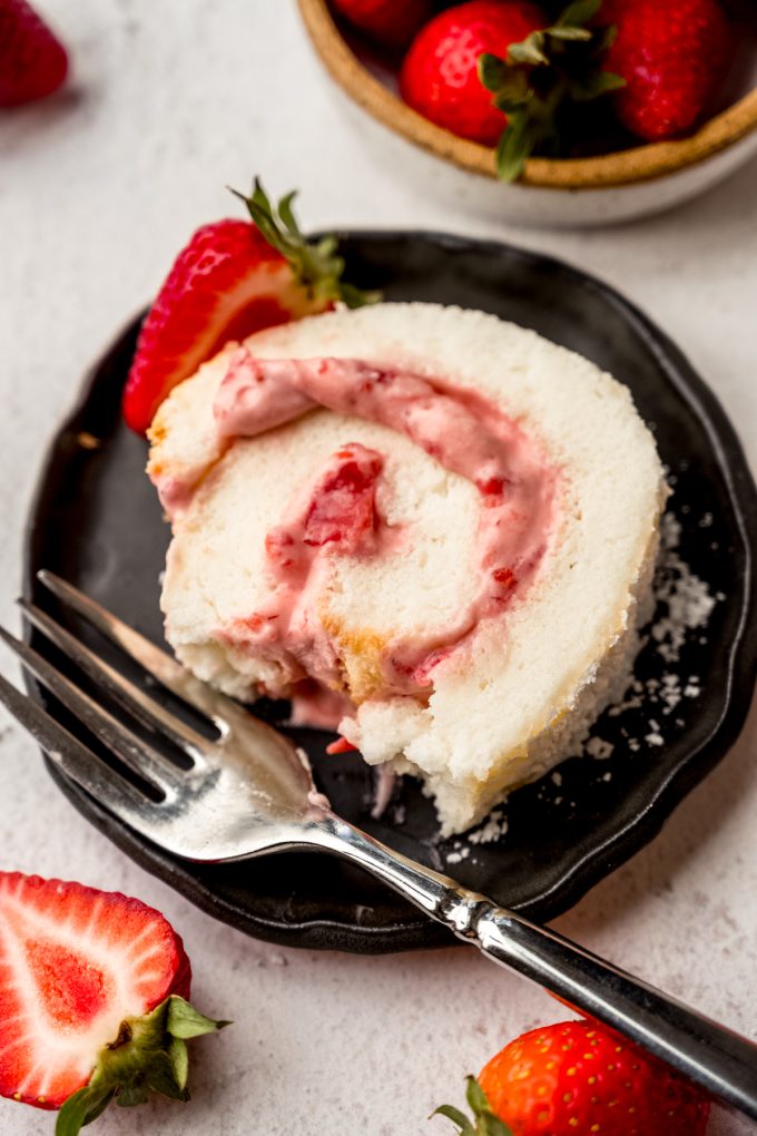 A slice of strawberry Swiss roll cake on a plate with a bite taken out of it.