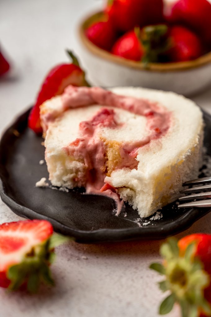 A slice of strawberry Swiss roll cake on a plate with a bite taken out of it.