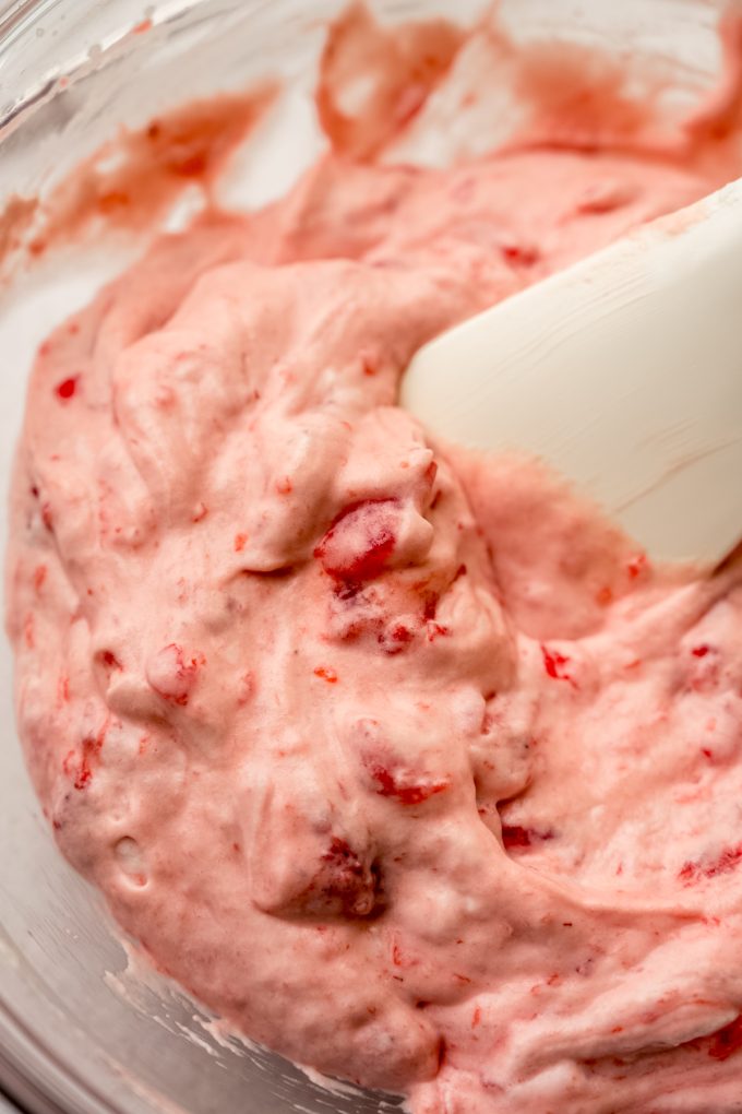 Someone is using a spatula to stir strawberry puree into whipped cream.