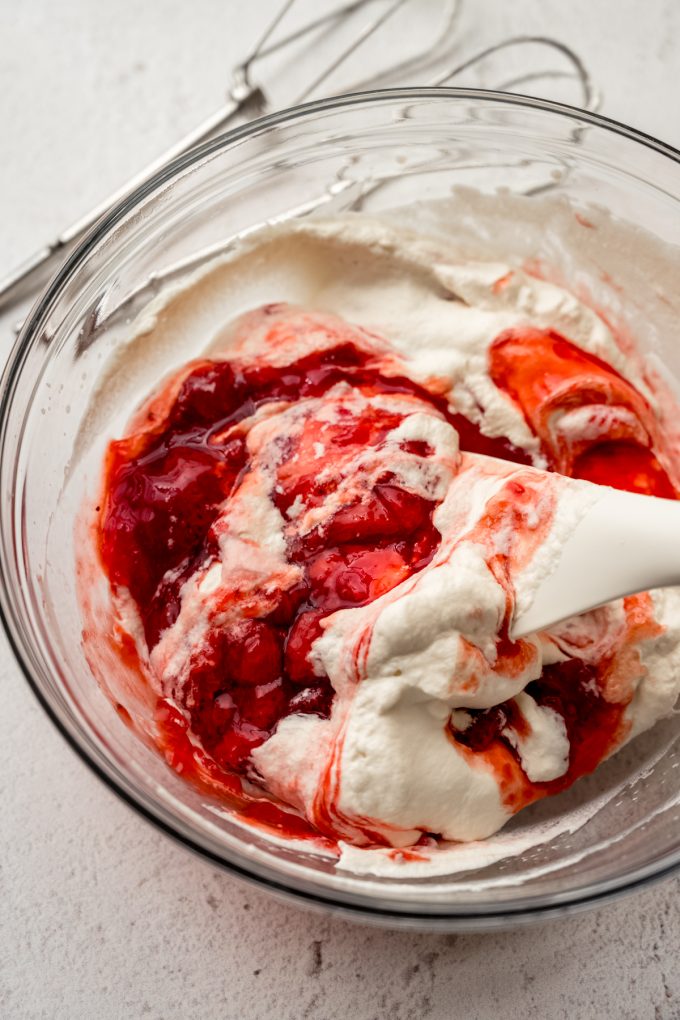 Someone is using a spatula to stir strawberry puree into whipped cream.