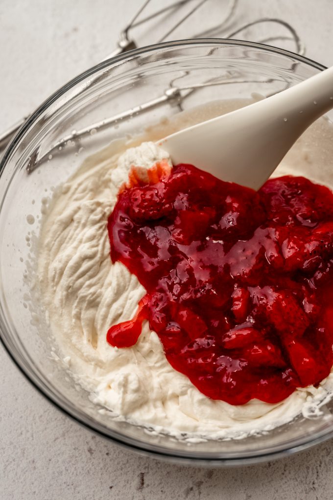 A bowl of whipped cream with strawberry puree and a spatula in it.