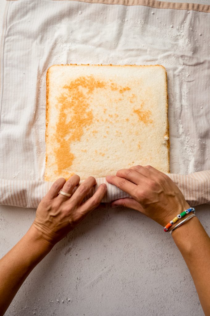 Someone is rolling up a layer of cake into a kitchen towel to make a Swiss roll cake.