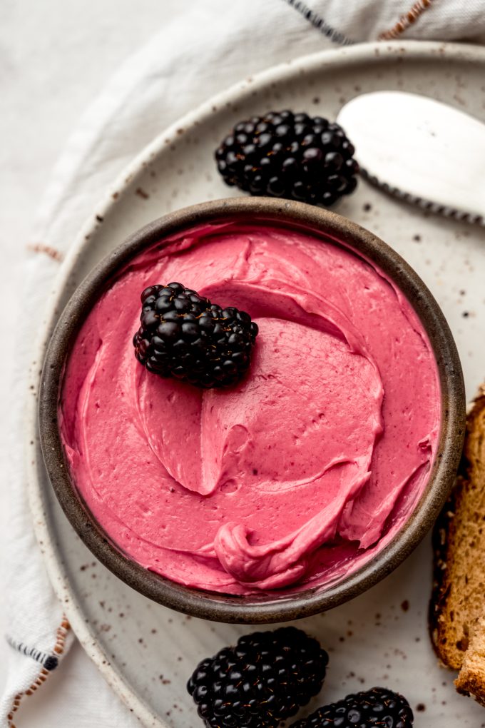 A small bowl of blackberry butter on a plate with a blackberry in it and other blackberries around the plate.