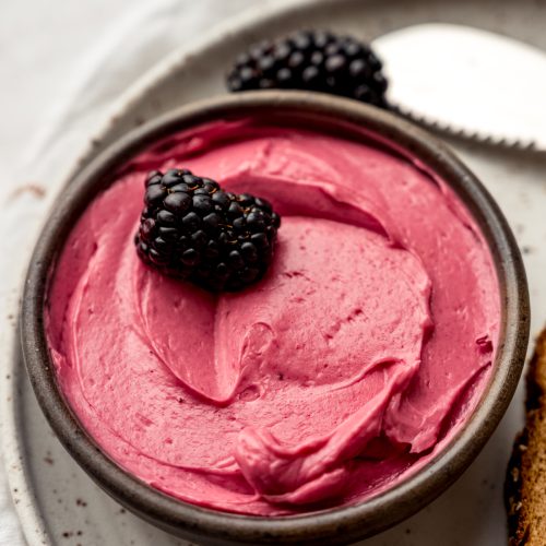 A small bowl of blackberry butter on a plate with a blackberry in it and other blackberries around the plate.