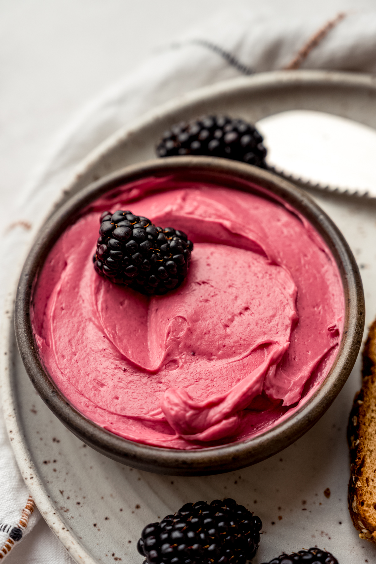 A small bowl of blackberry butter on a plate with a blackberry in it and other blackberries around the plate.