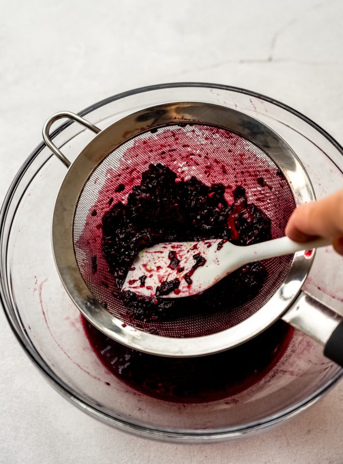 Someone is using a spatula to strain macerated blackberries through a mesh sieve.