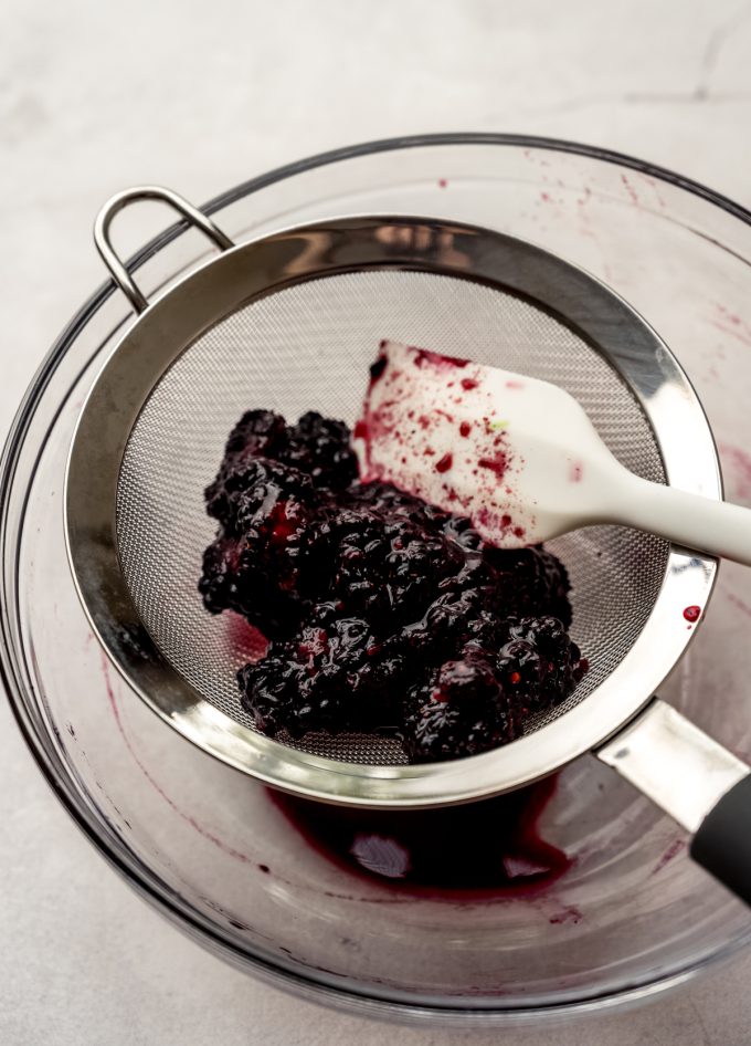 Someone is using a spatula to strain macerated blackberries through a mesh sieve.
