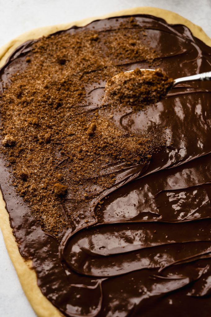 Someone is sprinkling cinnamon sugar onto a rectangle of dough spread with Nutella.