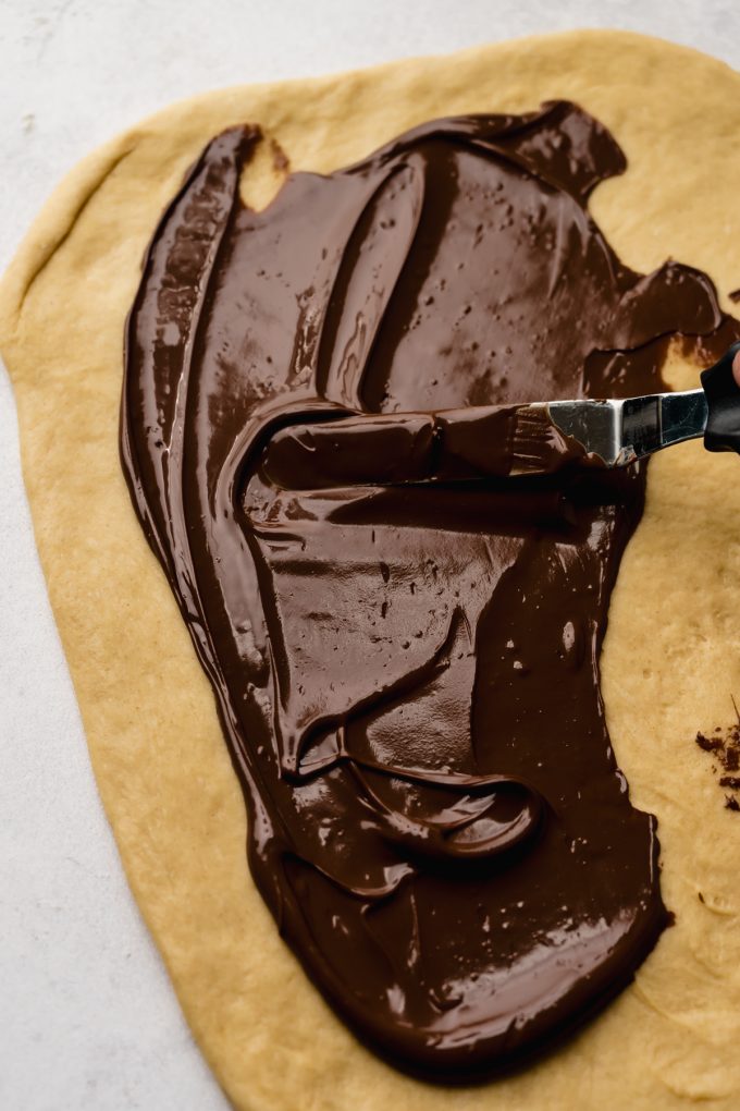 Someone is using an offset spatula to spread Nutella onto a rectangle of cinnamon roll dough.