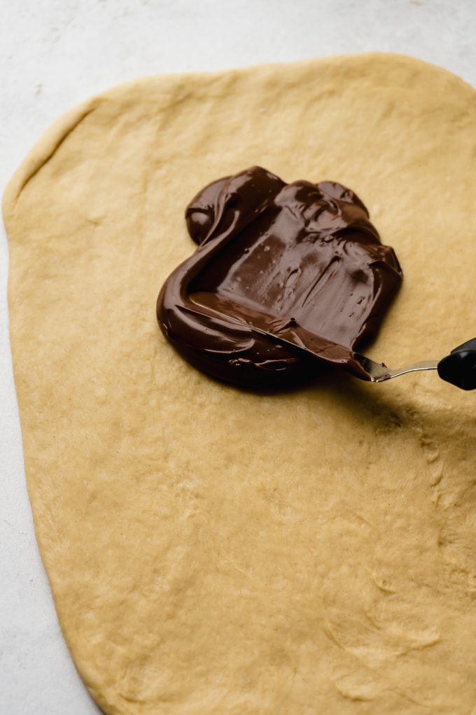 Someone is using an offset spatula to spread Nutella onto a rectangle of cinnamon roll dough.