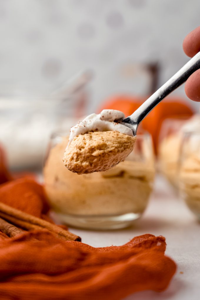 Someone is holding a small spoonful of pumpkin mousse close to the camera with a dessert cup of pumpkin mousse in the background.