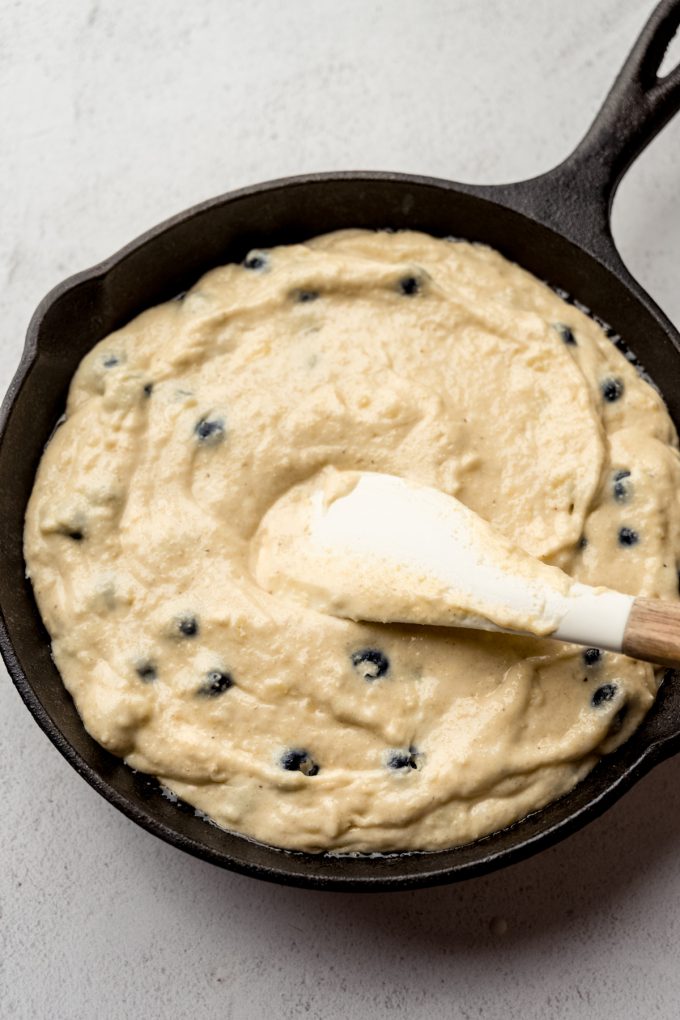 Someone is spreading blueberry cornbread batter into a cast iron skillet with a spatula.