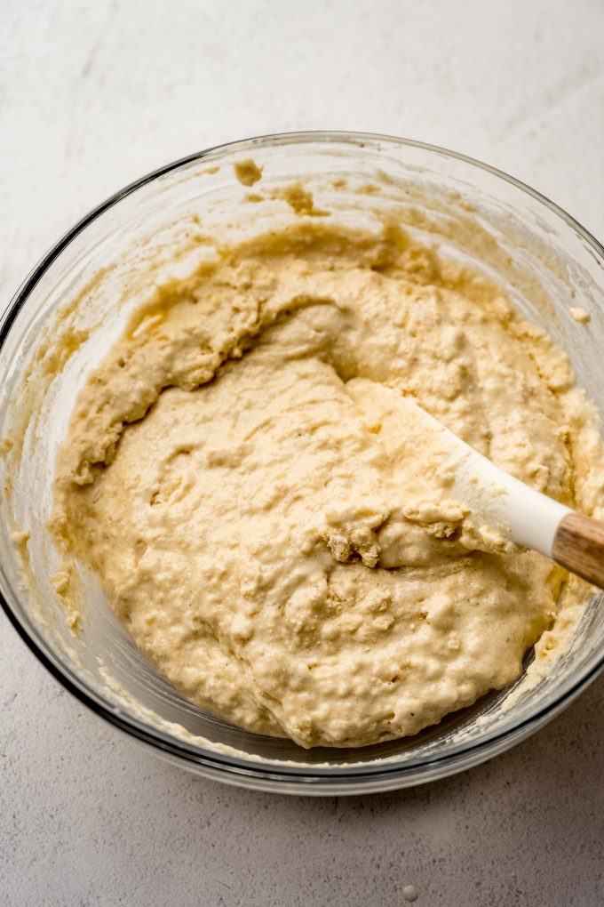 Someone is stirring cornbread batter in a large glass bowl with a spatula.