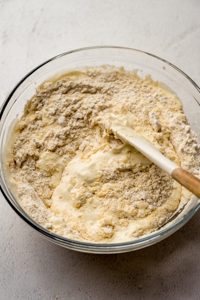 Someone is stirring cornbread batter in a large glass bowl with a spatula.