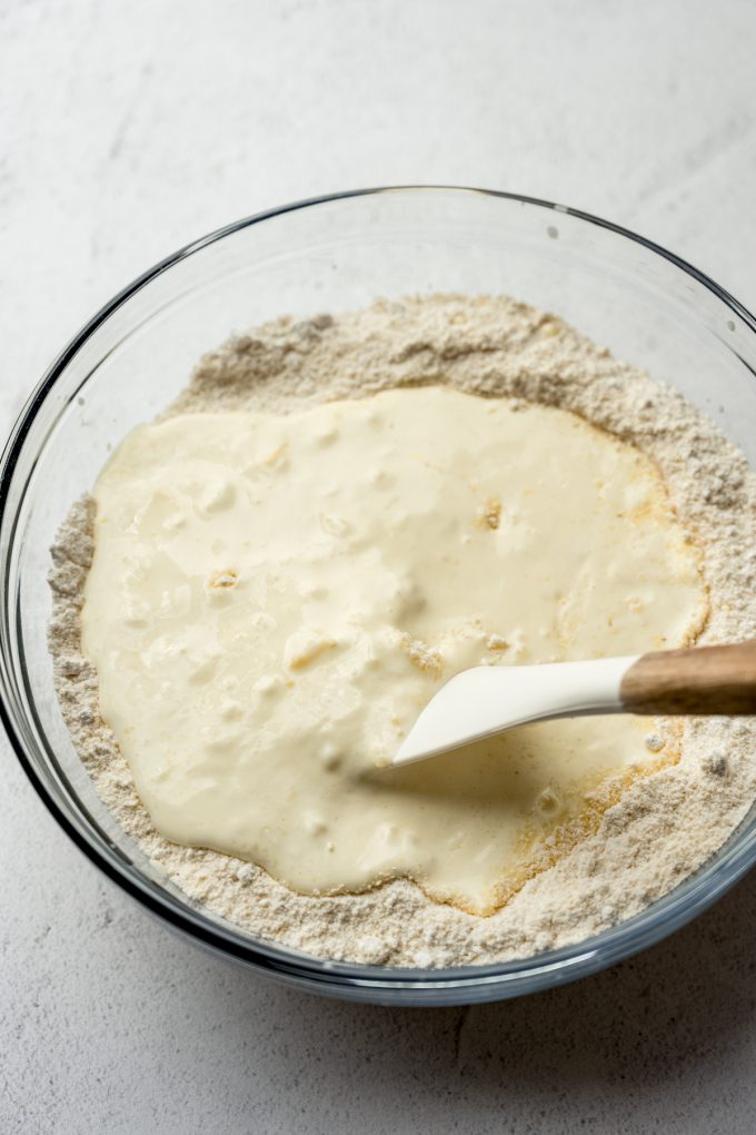 Someone is stirring cornbread batter in a large glass bowl with a spatula.