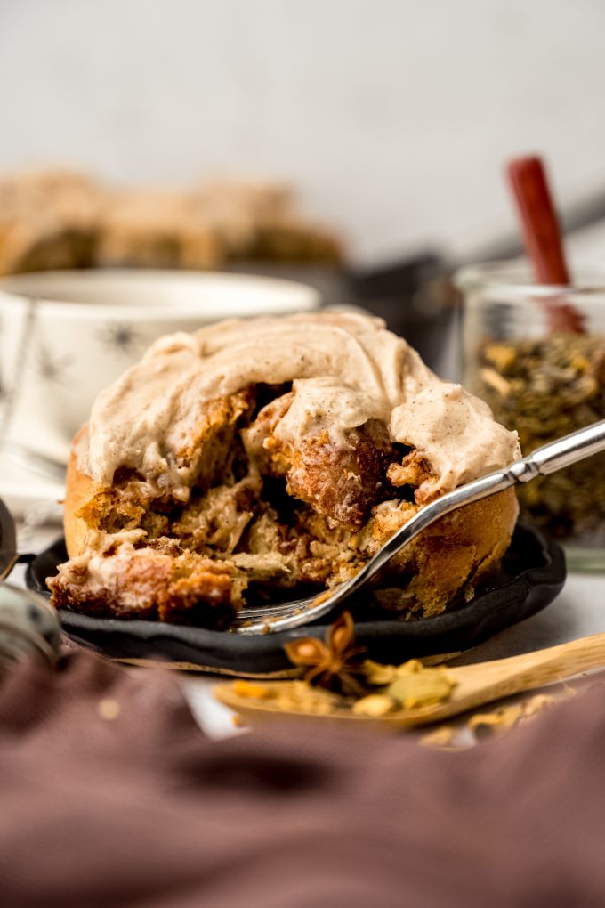 A chai cinnamon roll sitting on a plate with a fork and a bite has been taken out of it.