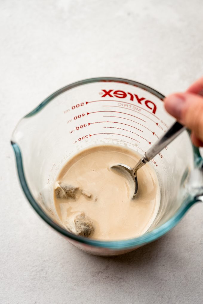 Someone is using a spoon to stir a measuring cup with milk and a chai bag in it.